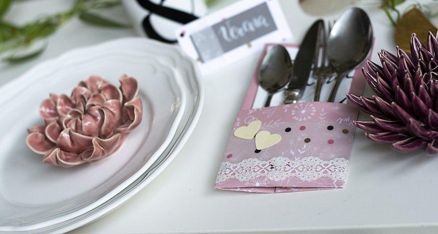 Gebastelte Bestecktasche aus rosa Papier mit Deko und Blumenteller auf weißem Tisch