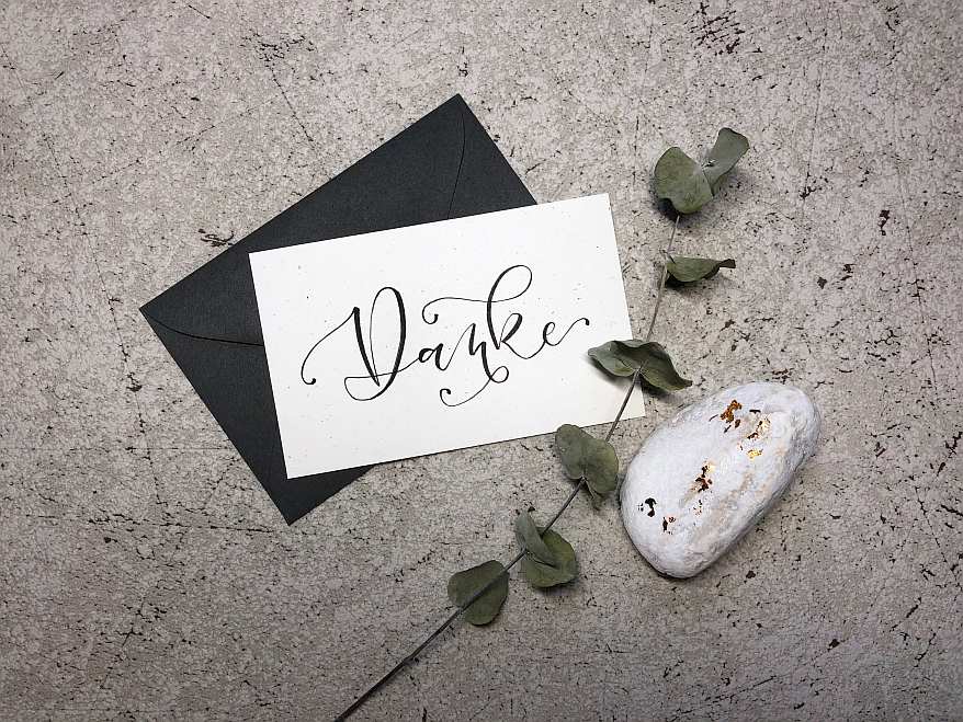 Auf einem Marmortisch liegt ein schwarzer Umschlag. Darauf liegt eine weiße Karte nmit einem schwarz gelettertem "Danke". Außerdem liegt als Deko ein Stein und ein Eukalyptusblatt daneben.