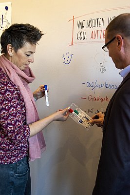 Zwei Mitarbeiter an einem Whiteboard reichen sich Whiteboard-Marker