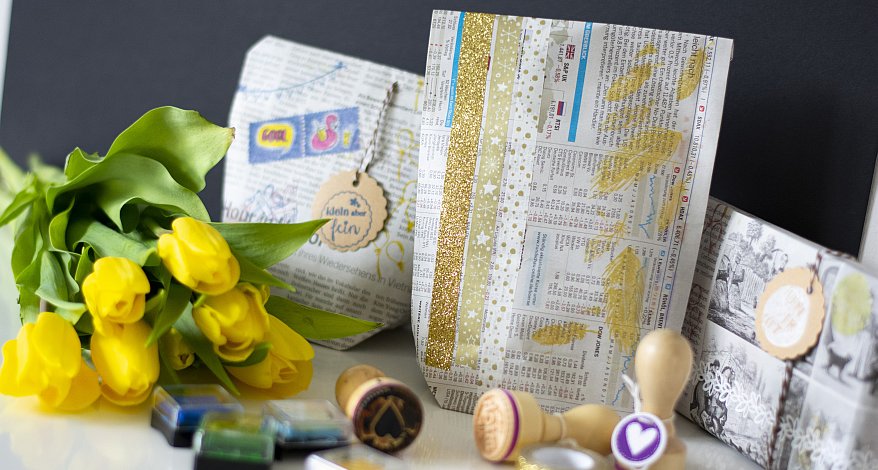 Auf einem weißen Tisch liegt ein Strauß gelber Tulpen. Daneben liegen drei Geschenke, die in Zeitungspapier eingepackt wurden und mit Stickern, Stempeln und Washitape verziert wurden. Im Vordergrund liegen einige Holzstempel und Stempelkissen.