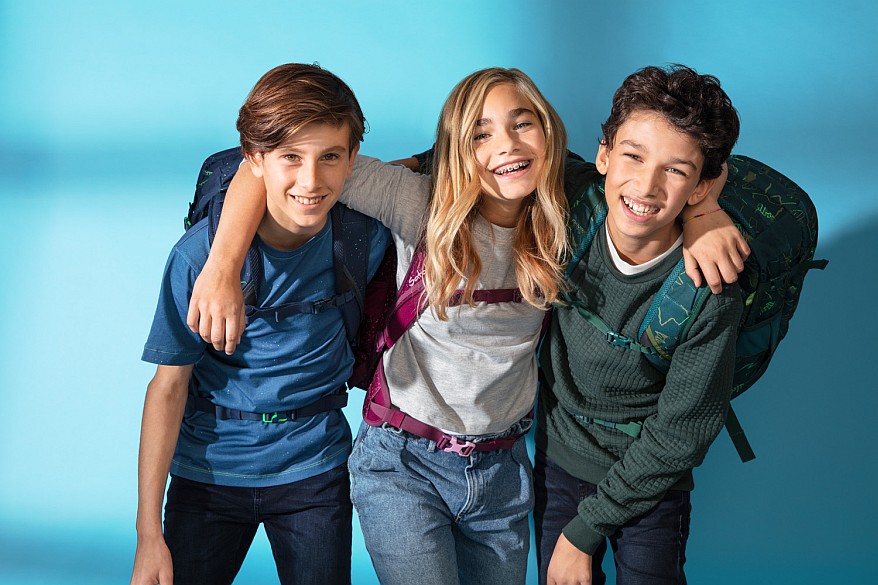 Drei Jugendliche mit Rucksäcken lachen und halten sich im Arm. Auf dem Rücken tragen sie Rucksäcke von satch.