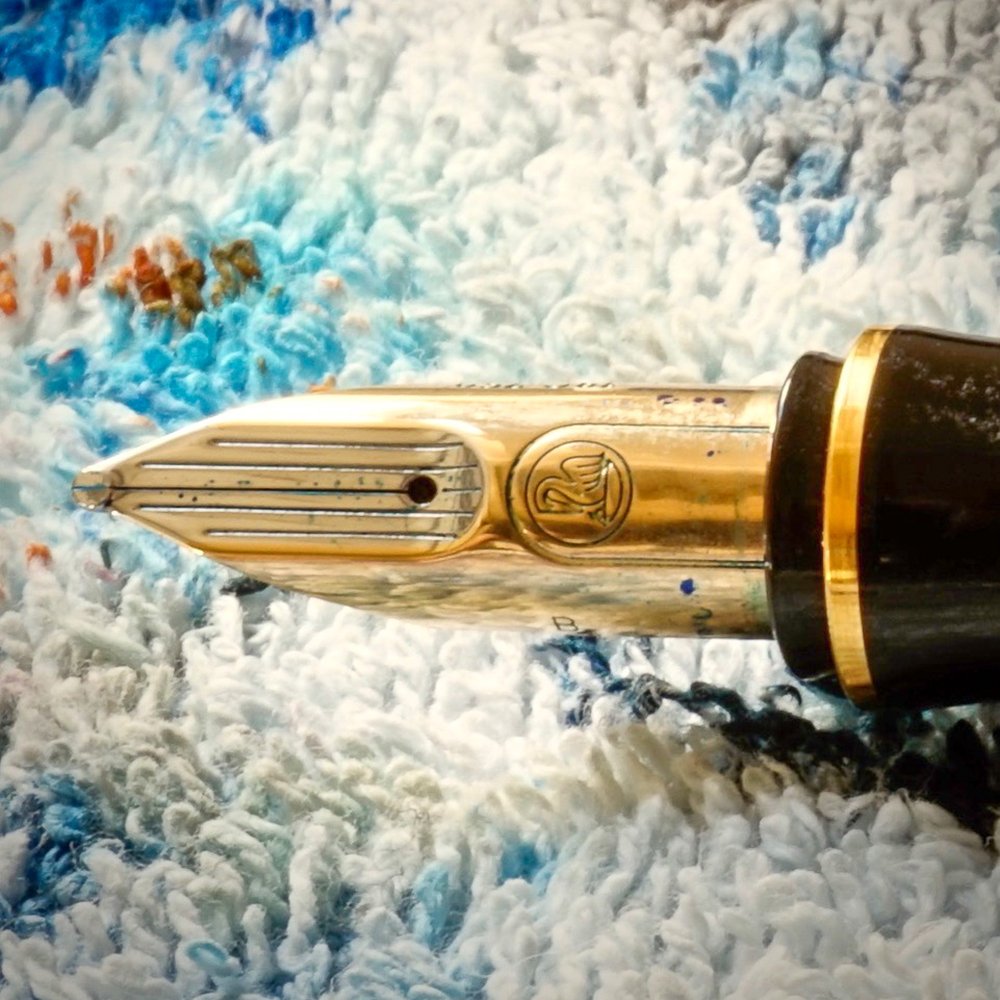 Ein Füller von Pelikan in der Nahaufnahme. Man sieht die hochwertige Goldfeder, an der etwas blaue Tinte haftet und das markante Logo von Pelikan.