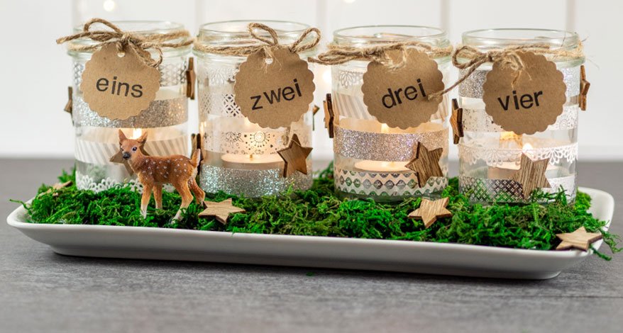 Ein selbstgebastelter Adventskranz mir vier Teelichtern in Einmachgläsern. Diese stehen auf Moos und wurden mit Washitape und Stickern beklebt. Um die Gläser ist mit brauner Schnur ein wellenförmiger Kreis aus Karton gebunden. Auf diese wurden die Zahlen eins bis vier gestempelt.