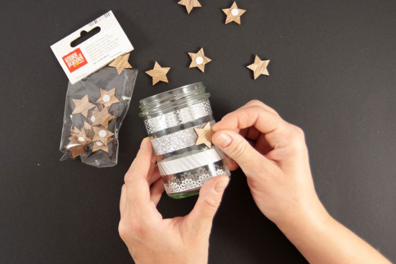 Ein mit weiß-grauem Washitape beklebtes Marmeladenglas wird mit Holzstern der Marke Knorr Prandell beklebt. Um das Glas liegen weitere Holzsterne verstreut. Der Untergrund ist schwarz.