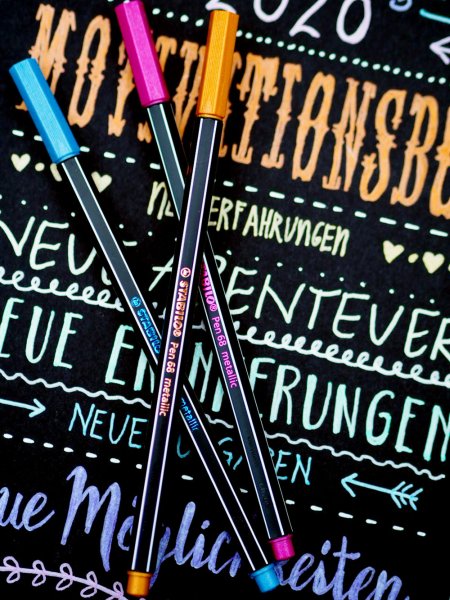 Ein schwarzer Karton wurde mit den Metallicmarkern von Stabilo belettert. Auf dem Karton liegen Metallic Marker in den Farben rosa, gold und blau.