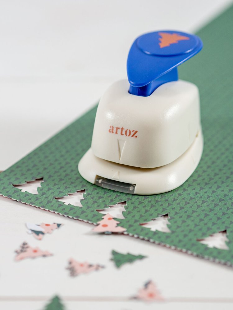 Auf einem grünen Motivpapier steht ein Motivstanzer der Marke Artoz. Aus dem Papier wurden bereits zahlreiche Tannenbäume ausgestochen, die darum verteilt liegen.
