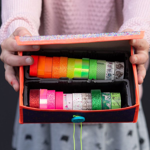 Kind hält bunte DIY-Box mit Glitzerdeckel und Knopfverschluss in Händen, darin bunte Washi-Tapes.