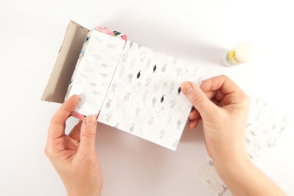 Ein Pappkarton wird mit weißen Motivkarton mit Fischen darauf beklebt. Im Hintergrund ist unscharf ein geöffneter Klebestift und weiterer Motivkarton zu sehen.