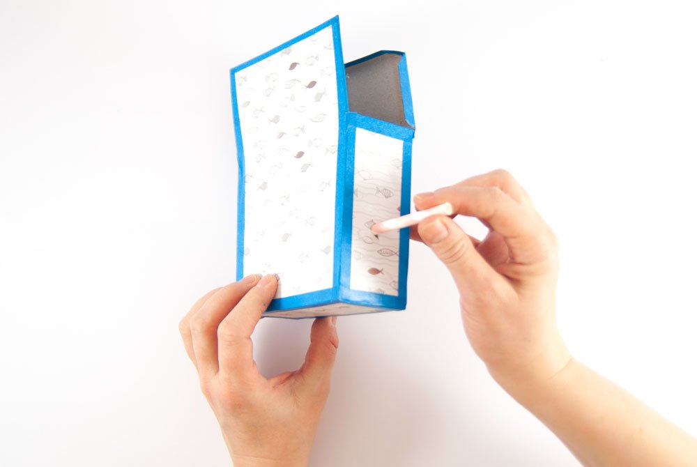 Eine mit Motivkarton und blauen Klebeband verzierte Papschachtel wird in einer Hand gehalten. Mit Hilfe eines Bleistiftes wird in die linke Seite der Schachtel ein Loch gebohrt.