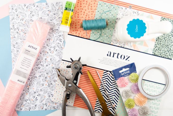 Bastelmaterialien wie buntes Papier, Deko-Aufkleber, Stanzer, Kordel, Krepp-Papier der Marke Artoz liegen auf einem Tisch.