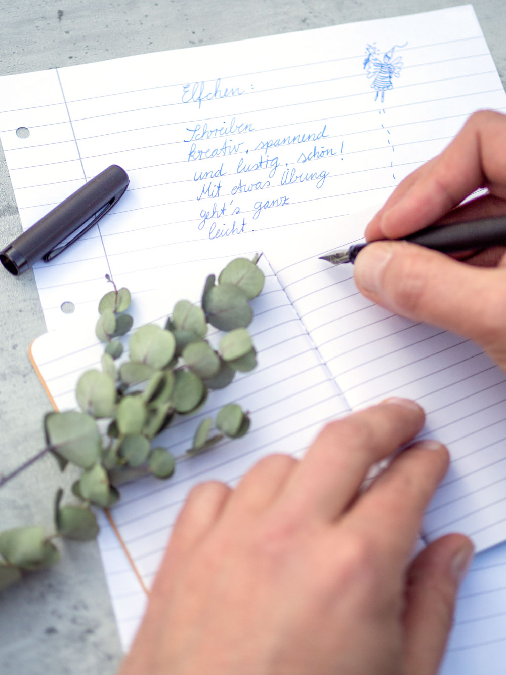 Hand schreibt mit Füllhalter ein Elfchen auf liniertes Papier, daneben Eukalyptuszweig.