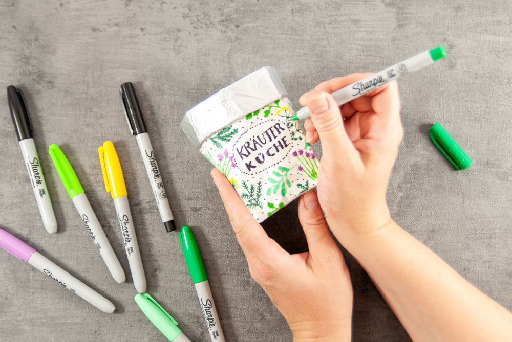 Auf einen Getränkekarton werden bunte Motive und das Wort Kräuertopf gelettert