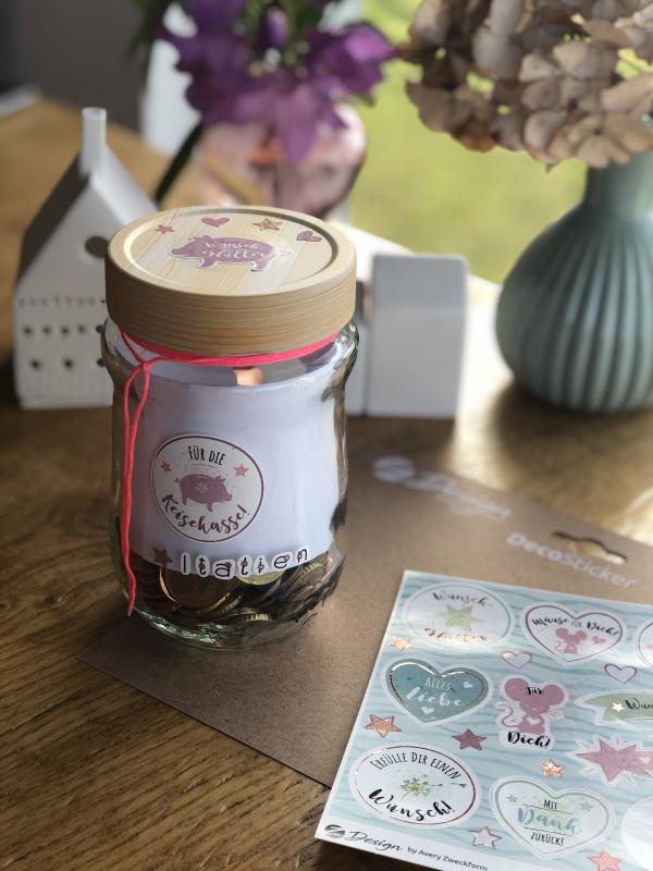 Ein Marmeladenglas wurde mit verschiedenen Stickern beklebt. In dem Glas ist Kleingeld. Am Bildrand sind Sticker, eine Vase und Deko zu sehen.