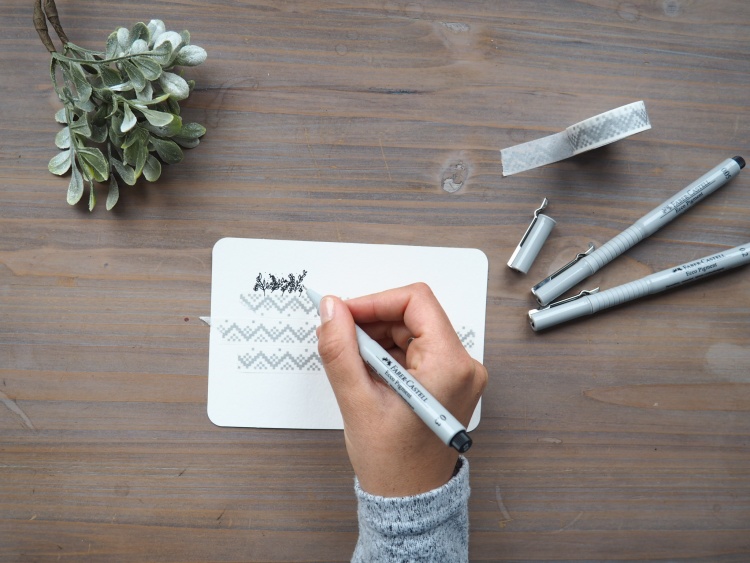 Eine Hand zeichnet einen Schriftzug auf eine Karte, die mit Washitape beklebt ist.