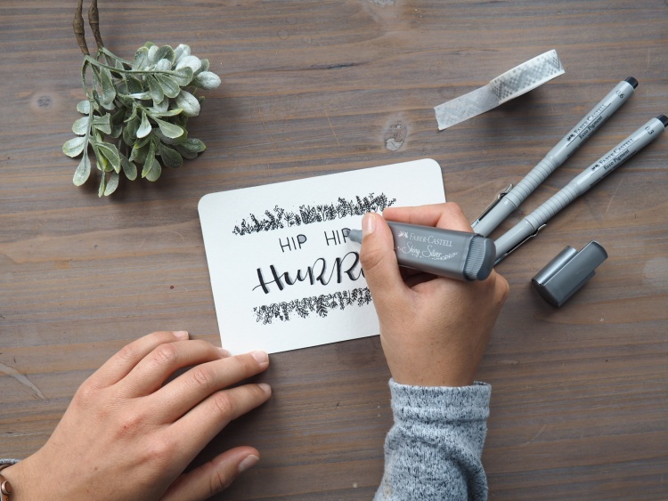Eine Hand malt mit einem silbernen Marker den Schriftzug "Hip Hip Hurra" auf eine Karte.