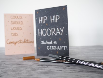 Eine beige und eine schwarze selbst beschriftete Karte stehen auf einem Tisch, davor 2 Stabilo Stifte.