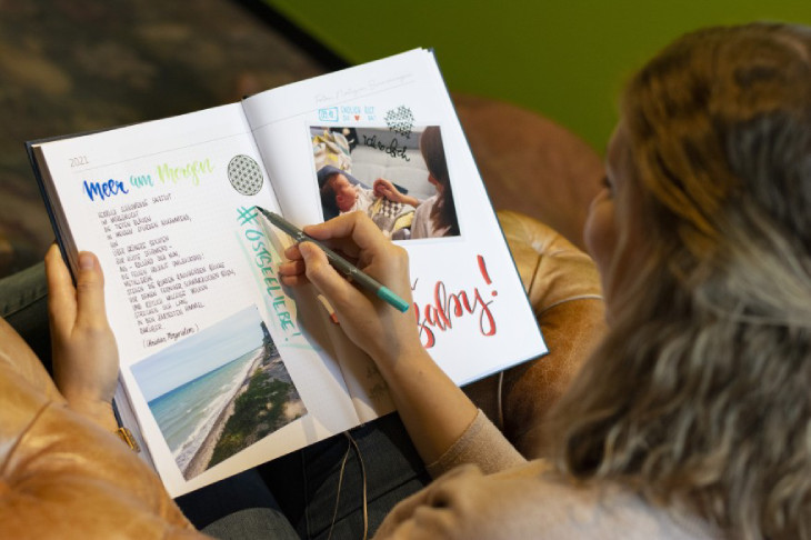 Eine junge Frau sitzt auf einer Ledercouch und halt den 10-Jahreskalender in der Hand. Auf einer Doppelseite hat sie Fotos eingeklebt und Texte in bunter Letteringschrift gestaltet. Mit einem türkisen Fineliner schreibt sie gerade etwas in den Kalender.