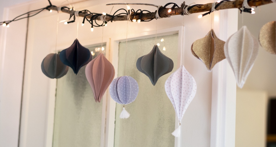 Eine aus Papier gebastelte Girlande als Tropfen und Kugeln in unterschiedlichen Farben hängen an einem beleuchteten Ast vor einem Fenster.