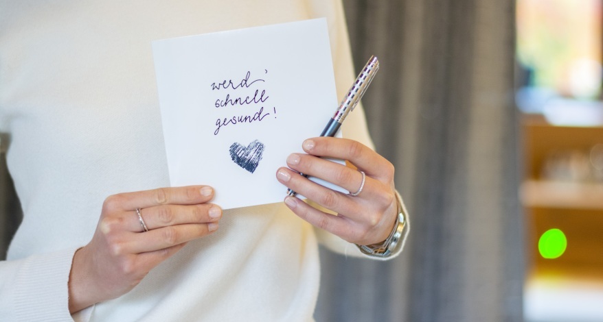 Eine Frau hält eine selbst gestaltete Gute Besserung Karte mit einem Stift in der Hand in die Kamera.