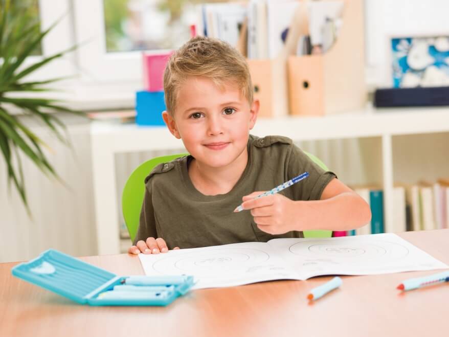 Ein Junge hält einen Griffix Bleistift von Pelikan in der rechten Hand und lächelt in die Kamera.Vor ihm liegen weitere Stidte, eine Stiftebox und ein Übungsblatt zum Schreibenlernen. Der Junge sitzt an einen Schreibtisch aus Holz. Im Hintergrund ist eine Pflanze sowie ein Regal mit Büchern und Zeitschriften zu erkennen.