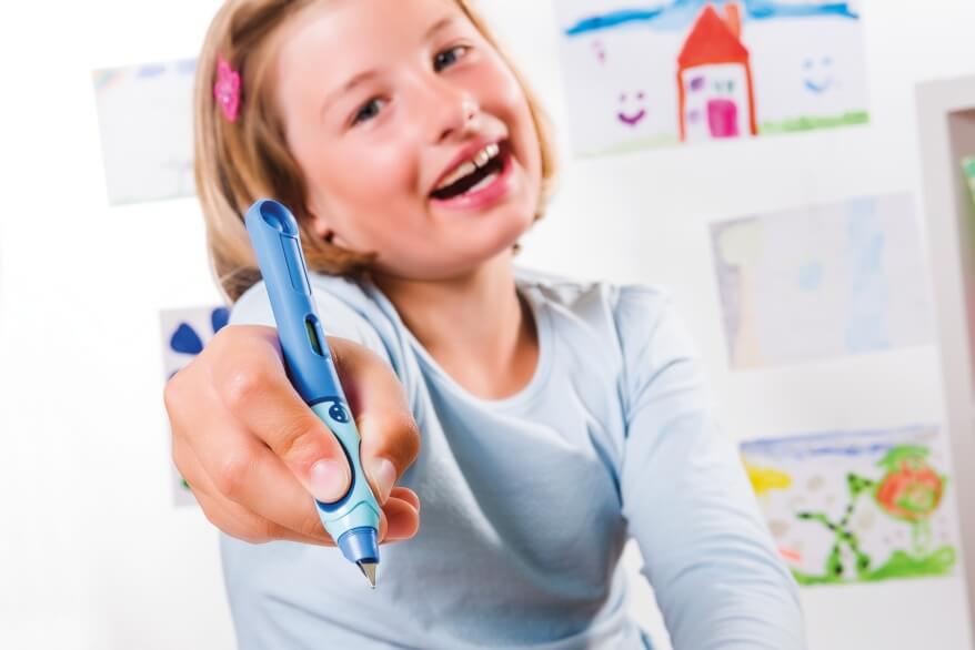 Ein Mädchen im Grundschulalter hält einen blauen Schreiblern-Füller von Pelikan in die Kamera und lacht dabei. Sie trägt ein hellblaues Longsleeve und eine rosa Spange im blonden Haar. Im Hintergrund sind selbstgemalte Bilder zu sehen.