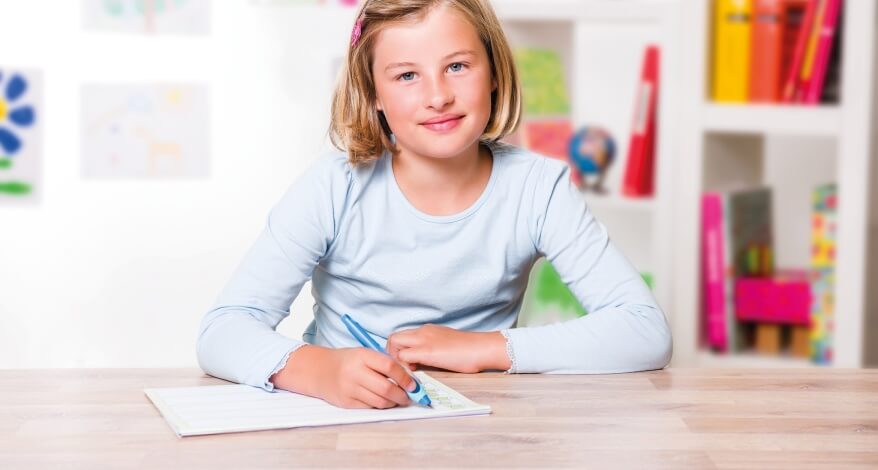 Ein Mädchen im Grundschulalter hält einen blauen Schreiblern-Füller von Pelikan in die Kamera und lacht dabei. Sie trägt ein hellblaues Longsleeve und eine rosa Spange im blonden Haar. Im Hintergrund sind selbstgemalte Bilder zu sehen