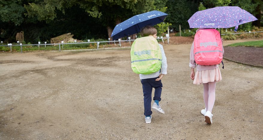 Zwei Kinder gehen mit aufgespannten Regenschirmen über einen Weg. Sie tragen zwei Schulranzen von ergobag, die mit Regencapes in Neonfarben geschützt sind.