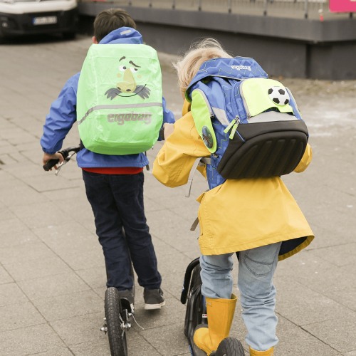 Zwei Kinder in Regenkleidung fahren auf Rollern über einen gehweg. Sie tragen beide Schulranzen von ergobag.