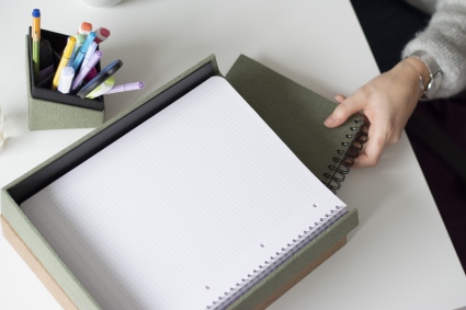 Hand zieht im Homeoffice ein Notizbuch aus S.O.H.O.-Ablageschale von Rössler. Daneben steht ein Stifteköcher