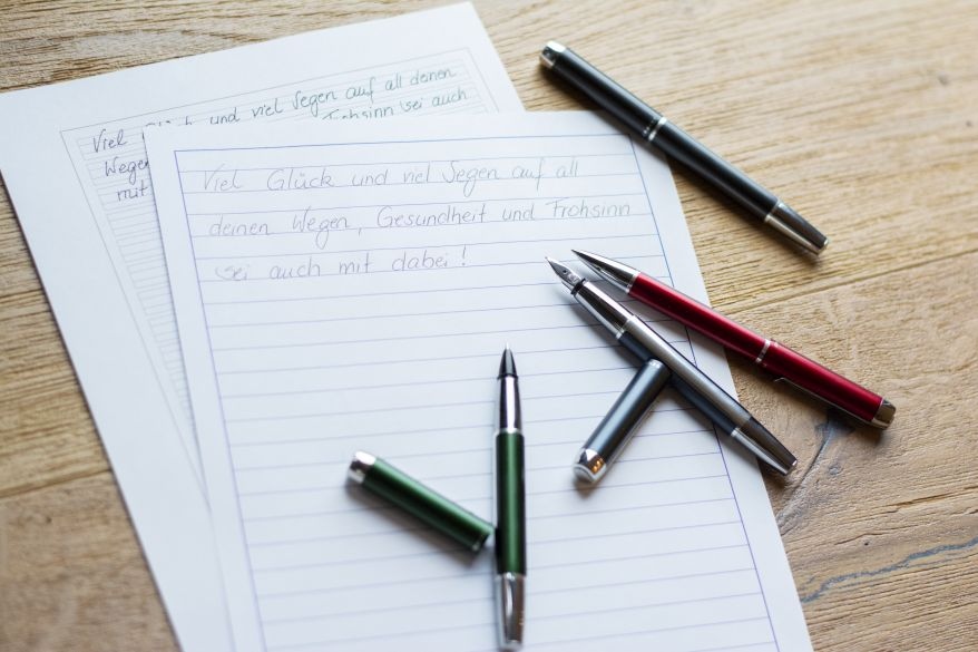 Auf zwei Blättern Papier mit verschiedenen Lineaturen steht geschrieben "Viel Glück und viel Segen auf all deinen Wegen, Gesundheit und Frohsinn sei auch mit dabei" als Übung für die Schönschrift. Verschiedene Stifte und Schreibgeräte der Serie Pura von Pelikan liegen auf dem Papier.