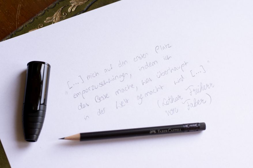 Auf einem Blatt ist ein Zitat von Lothar von Faber mit Bleistift geschrieben. Auf dem Blatt liegt ein schwarzer Bleistift von Faber-Castell