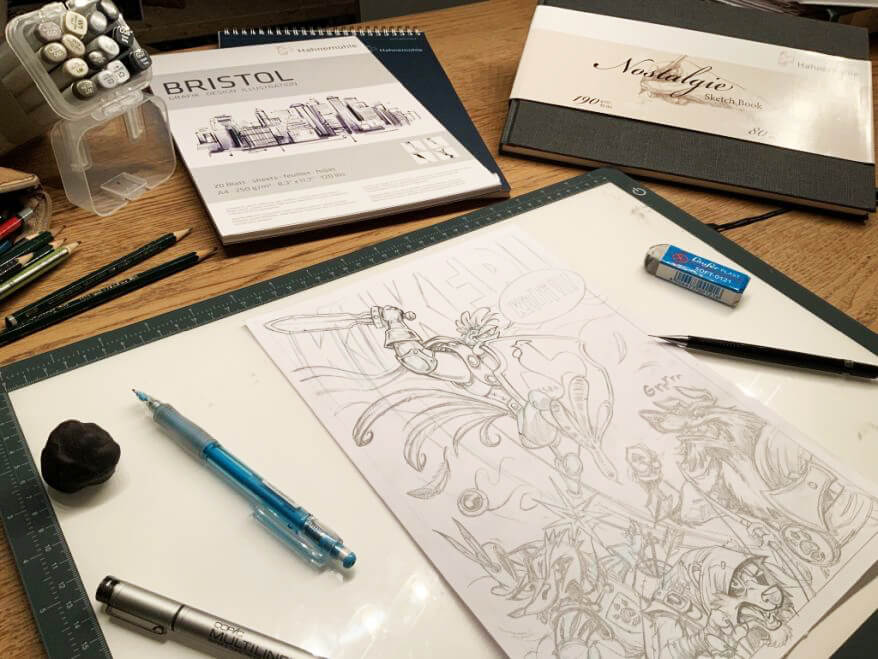 Blick auf den Schreibtisch eines Comiczeichners: Man sieht eine Bleistiftskizze auf einem Leuchttisch liegen, drumherum Stifte und Papiere zum Zeichnen, einen Leuchttisch sowie ein Skizzenbuch und Bristolpapier von Hahnemühle.