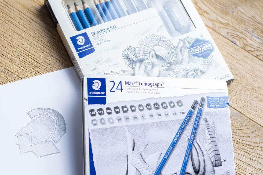 Zwei Zeichensets der Marke Staedtler liegen auf einem Schreibtisch, daneben ist das Logo von Staedtler auf einem Blatt Papier nachgezeichnet.