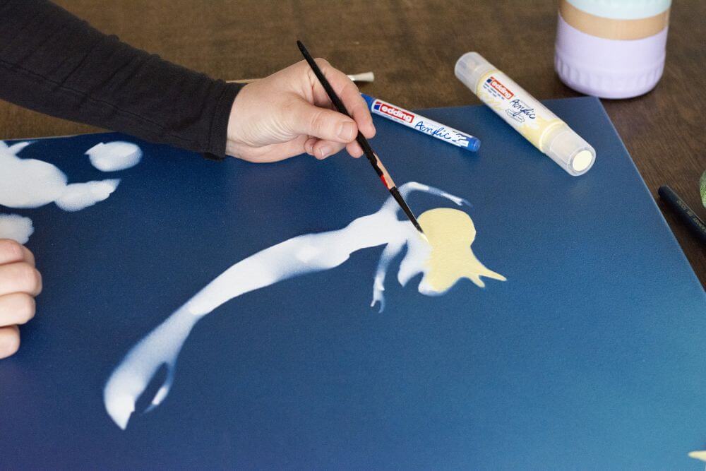 Eine Hand malt mit Pinsel und Acrylfarbe eine Meerjungfrau auf Leinwand.