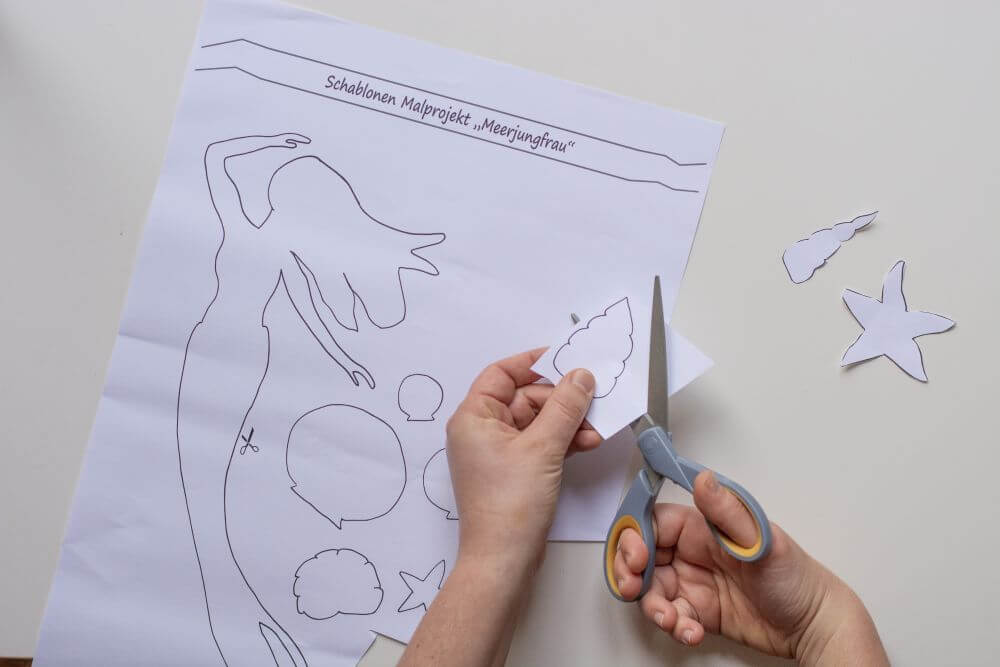 Hände schneiden mit einer Schere Schablonen aus Papier in Form einer Meerjungfrau und Muscheln aus.