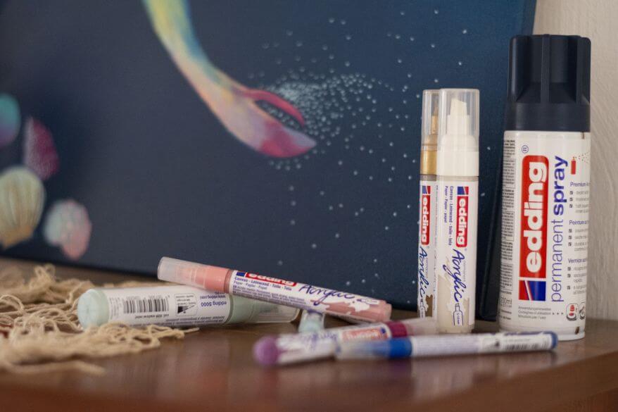 Sprühfarben und Acrylmarker von Edding stehen vor einer Leinwand mit gemalter Meerjungfrau.
