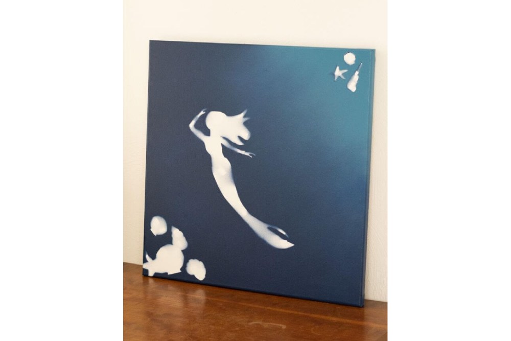 Eine Leinwand, die mit blauer Sprühfarbe besprüht wurde, steht auf einer Anrichte. Zu sehen sind die ausgesparten Konturen einer Meerjungfrau und Muscheln.