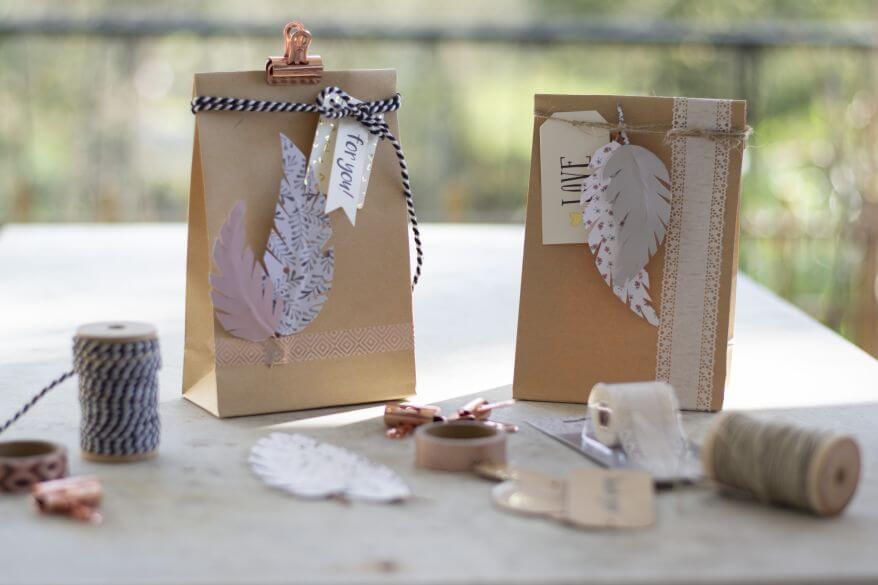 Auf einem Tisch stehen zwei Geschentüten, die mit selbstgemachter Deko gestaltet sind: Do it yourseld-Papierfedern, Dekoschnur, Washitape und Geschenkanhänger. Drumherum steht das Dekomaterial.