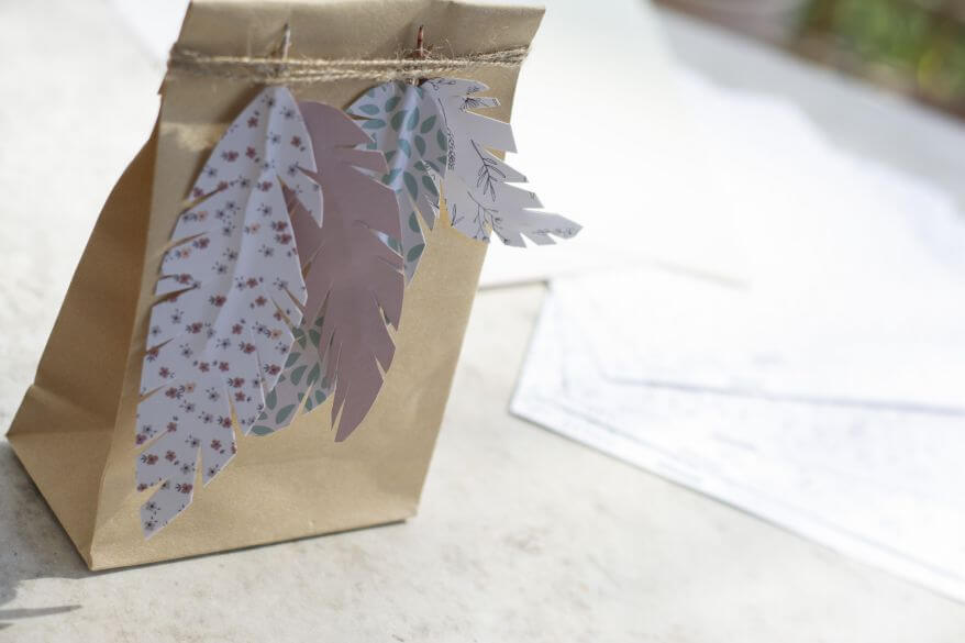 An einer Geschentüte hängen an einer Schnur verschiedene Do it yourself-Papierfedern in unterschiedlichen Farben und Formen.