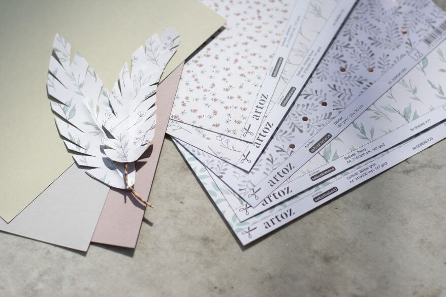 Mehrere selbstklebende Motivkartons und Papiere von Artoz liegen auf einem Tisch. Darauf liegen zwei Do it yourself-Papierfedern.