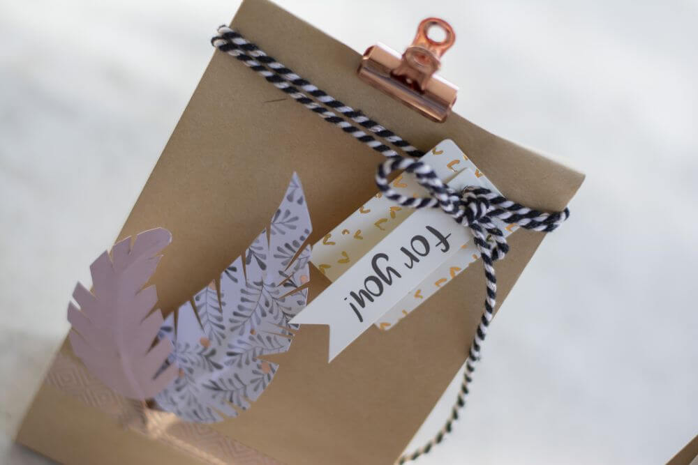 An einer Geschenktüte hängt ein Geschenkanhänger mit der Aufschrift "for you", eine schwarz-weiße Kordel und mit Maskingtape sind Do it yourself-Papierfedern an ihr befestigt. Ein Metallclip verschließt die Tüte.