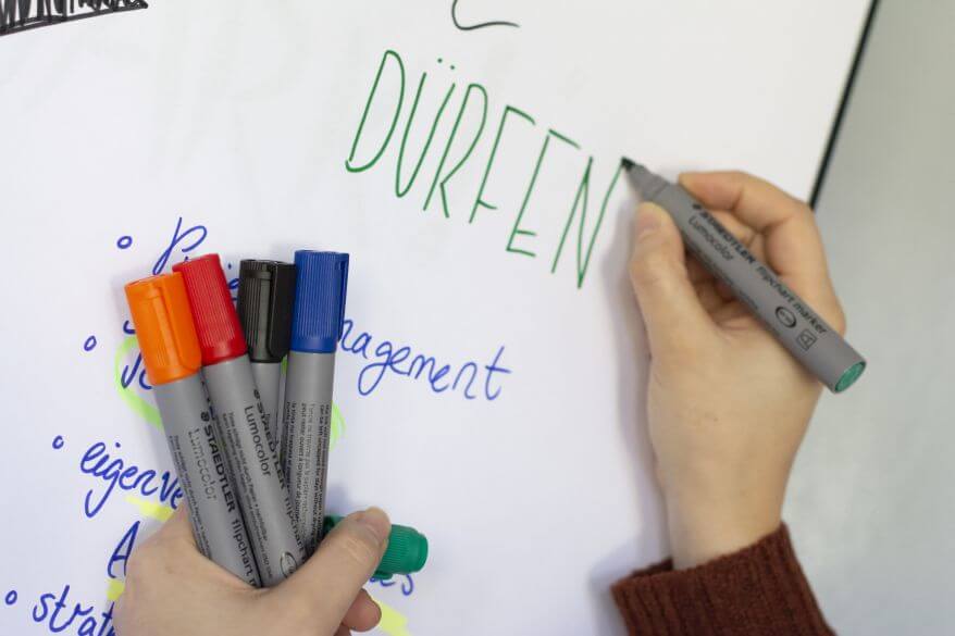 Eine Hand schreibt mit dem Flipchartmarker Lumocolor von Staedtler "Dürfen" auf ein Flipchartpapier. In der anderen Hand hält sie Flipchartmarker in weiteren Farben.