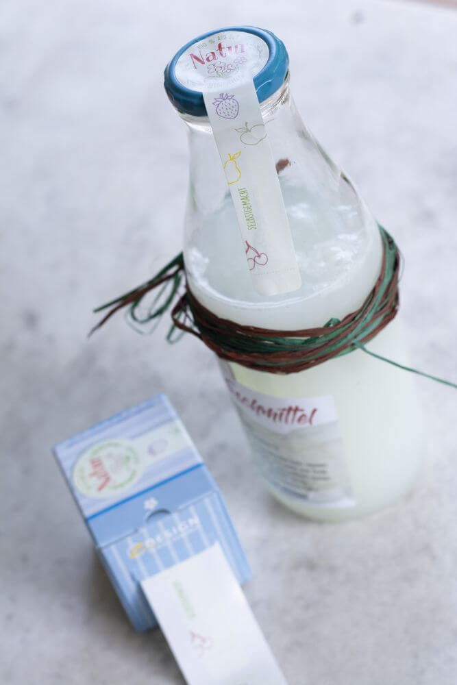 Eine Glasflasche mit selbstgemachtem DIY-Waschmittel steht auf einem Tisch, daneben Sticker von der Rolle von Avery Zweckform.