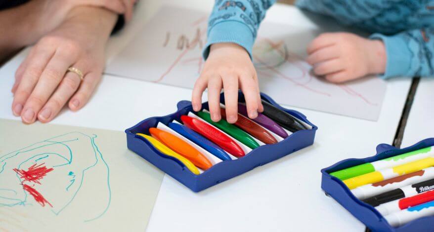 Eine Kinderhand greift zu bunten Wachsmalstiften, die auf dem Tisch liegen. Auf dem Tisch liegen bemalte Blätter.
