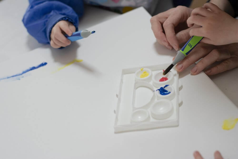 Ein Erwachsener und Kinder geben Gouachefarben aus der Kreativfabrik von Pelikan auf eine Farbpalette.