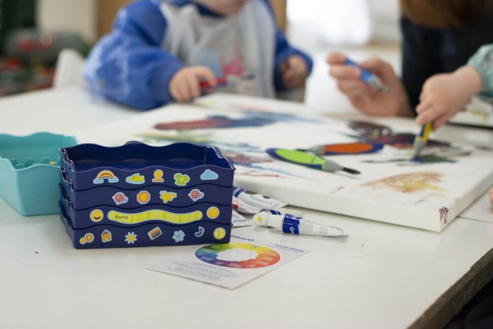 Im Vordergrund steht auf einem Tisch eine mit Stickern beklebte Kreativfabrik von Pelikan, im Hintergrund malen Kinder auf eine Leinwand.