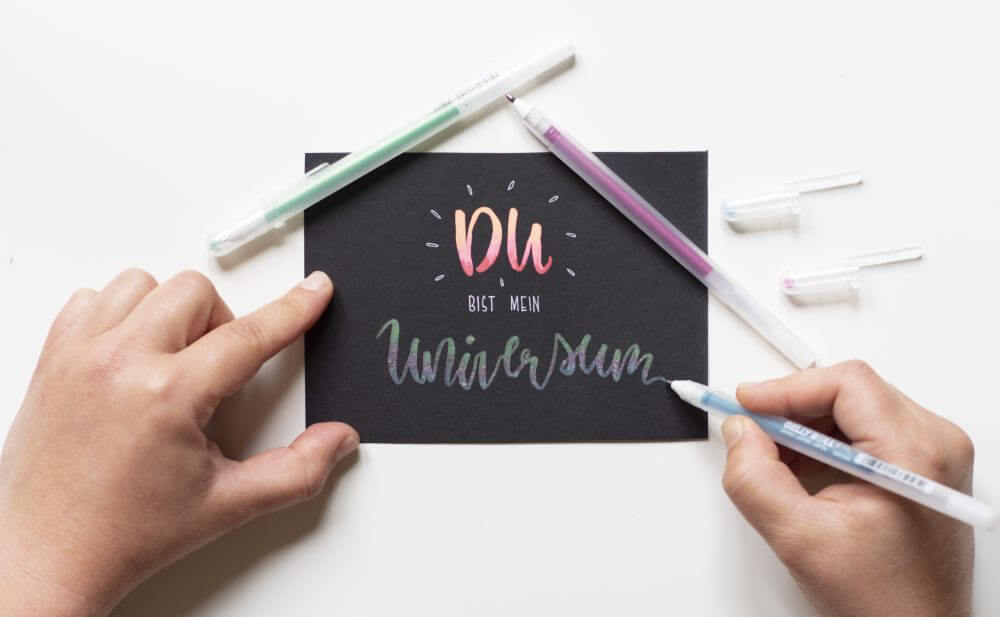 Anleitung Blending mit Gelstiften (Gelly Rolls von Sakura), Schritt 3: Mit Gelrollern in glitzernden Farben das Wort "Universum" mit Farbe und Farbverläufen füllen.