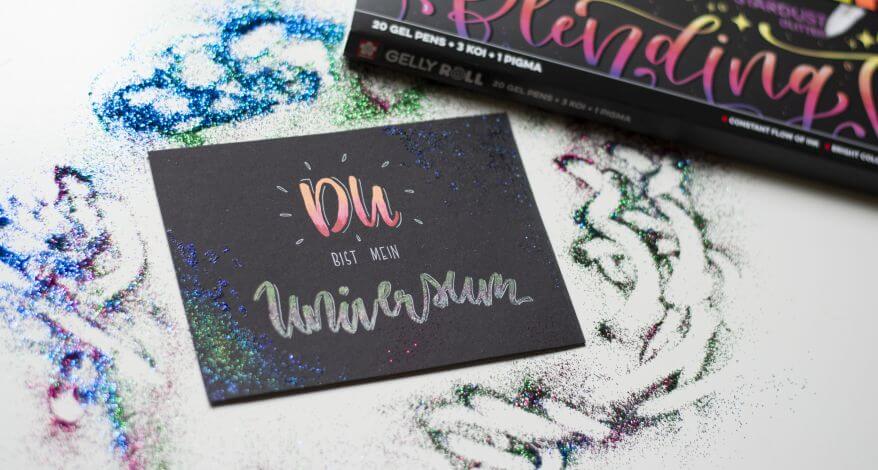 Eine selbstgemachtes Handlettering mit dem Schriftzug "Du bist mein Universum", der mit Gelstiften "Gelly Rolls" von Sakura gestaltet wurde, liegt auf einem Tisch. Das Papier ist dunkel, die Schrift leuchtet, drumerhum liegt Glitter verstreut.
