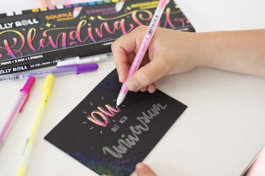 Eine Hand schreibt als Handlettering "Du bist mein Universum" mit Gelstiften "Gelly Rolls" von Sakura auf eine dunkle Karte. Im Hintergrund liegt das SAKURA Gelly Roll Frau Hölle Blending-Set, 24 Stifte.