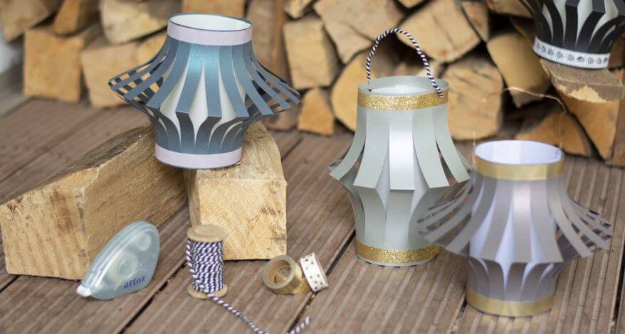 3 selbstgebastelte Papierlaternen stehen auf einer Terasse auf dem Boden vor einem Stapel Holz. Davor liegen Masking-Tape, Kordel und Kleberoller der Marke Artoz.